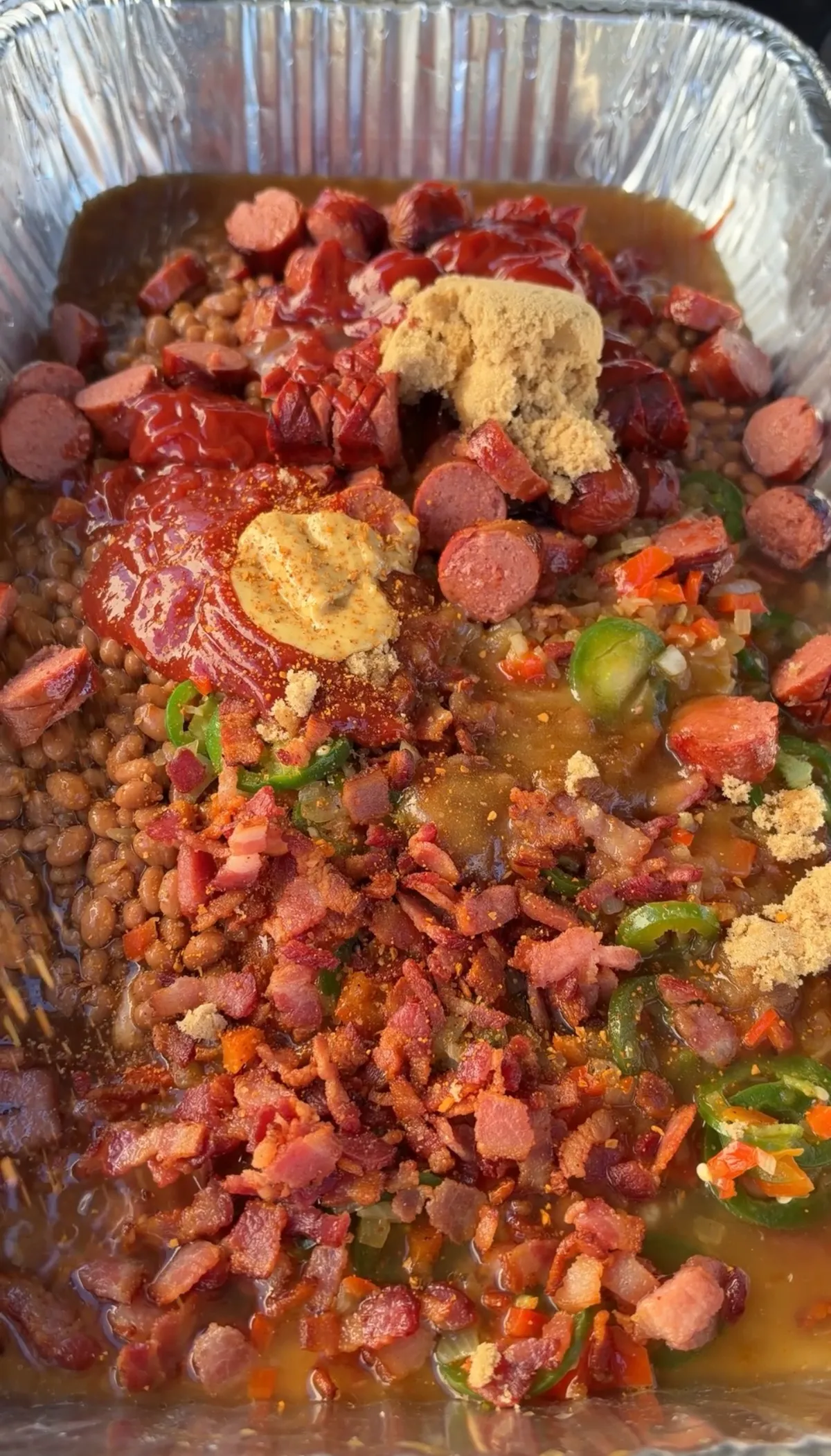 Foil pan with Bush's baked beans, sliced hot dog rounds, crispy bacon, jalapeños, brown sugar, ketchup, and dry mustard before smoking