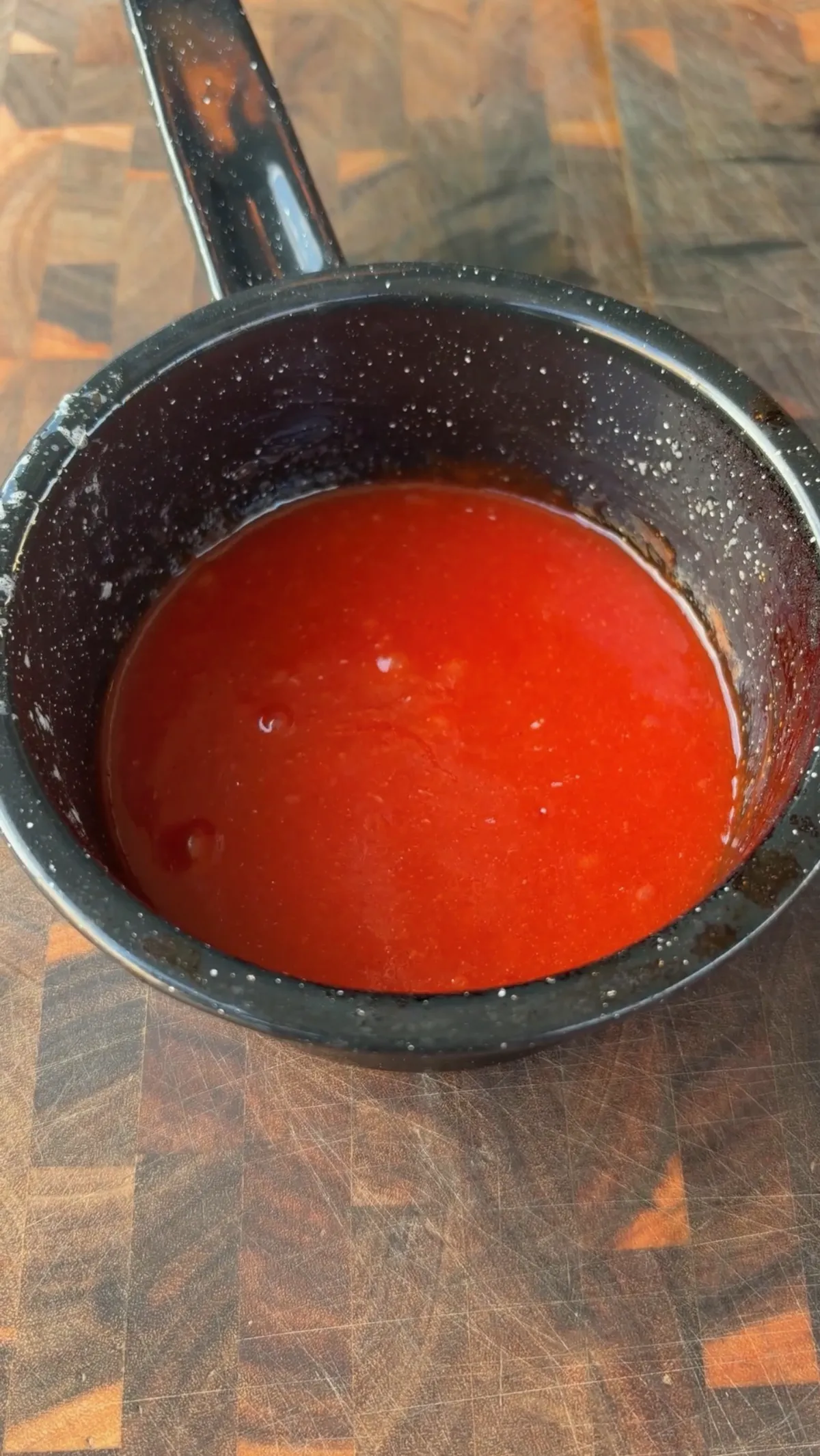 Red wing sauce injection mixture with butter, honey, and food coloring in a black saucepan on a wooden cutting board
