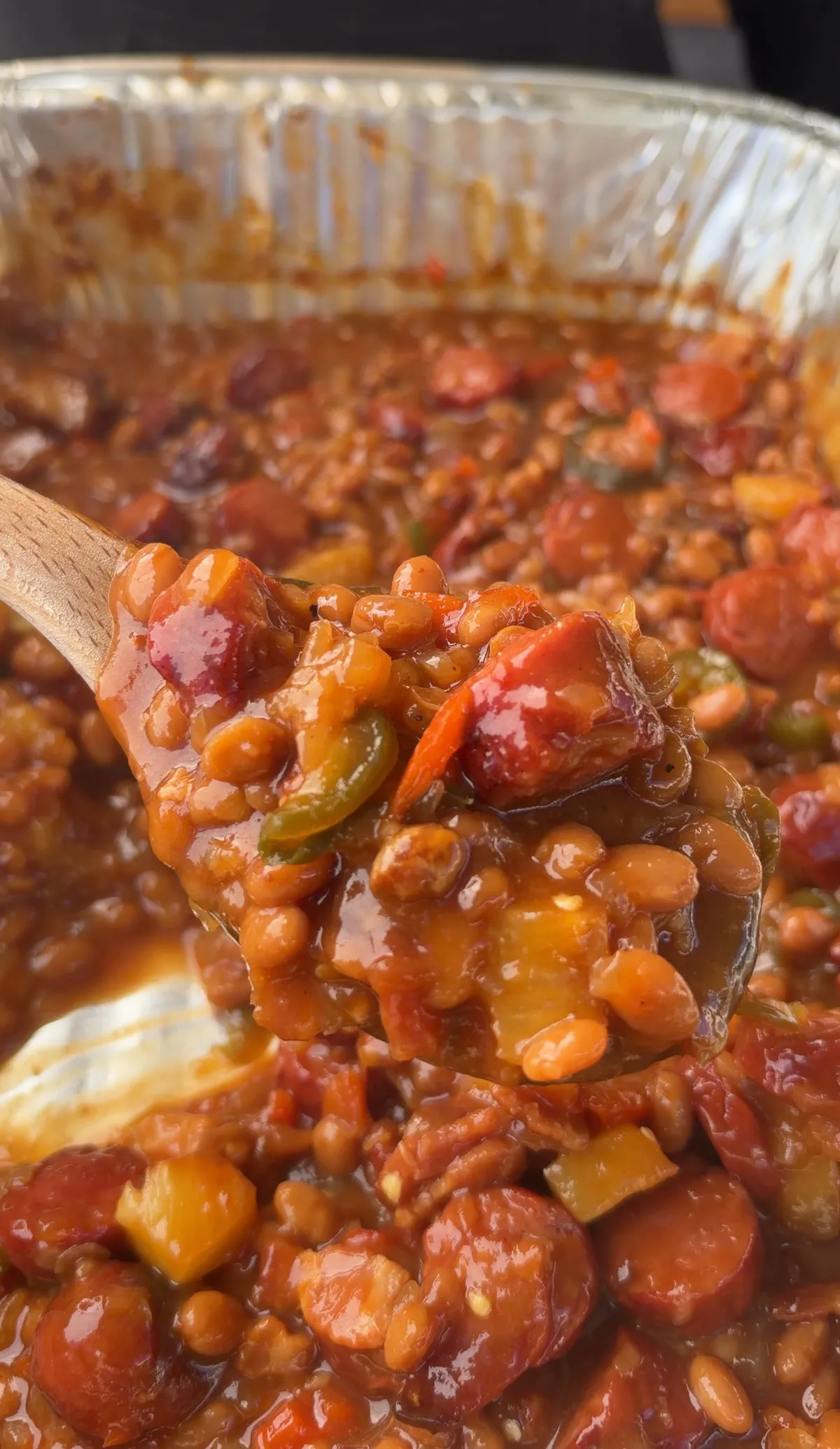 Wooden spoon lifting a scoop of smoked BBQ baked beans with hot dog rounds, jalapeños, and thick sauce from a foil pan