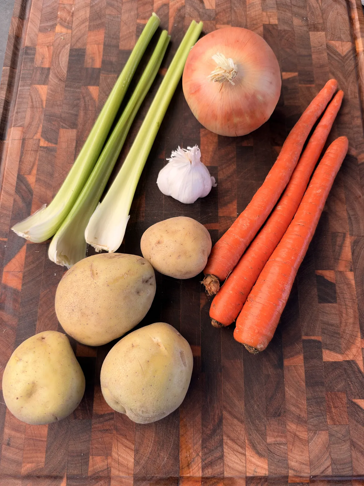 Yukon Gold potatoes, carrots, celery stalks, yellow onion, and garlic on a wooden cutting board